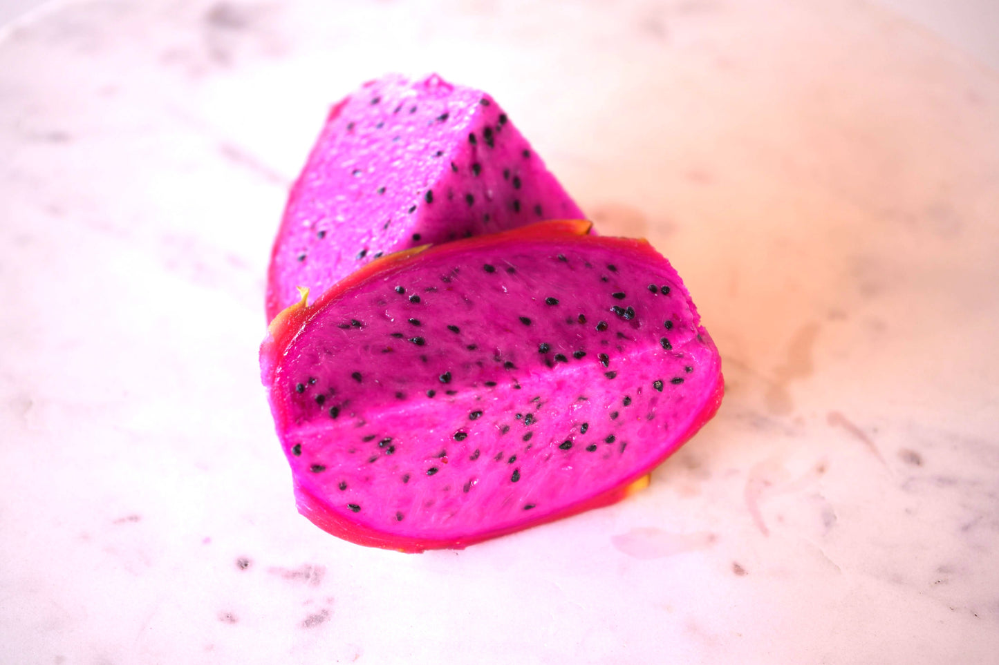 Two vibrant pink dragon fruit slices with black seeds on a light marble surface